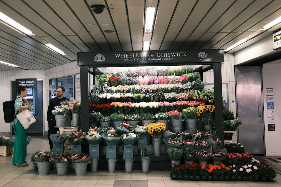 A floral display stand at Wheeler of Chiswick inside a London underground station, showcasing a variety of fresh flowers arranged in multiple tiers. The arrangement includes roses, tulips, sunflowers,