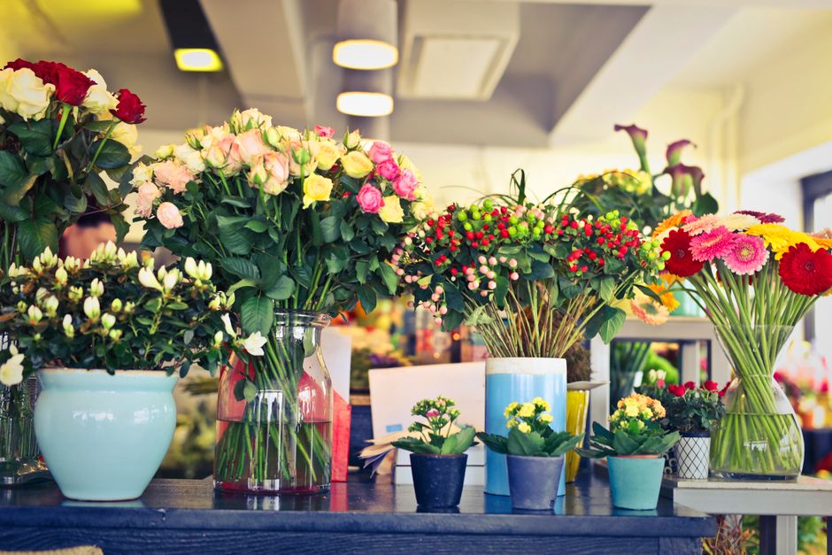 A variety of fresh floral arrangements displayed on a dark wooden table inside a florist shop. There are large bouquets of roses in soft pink, white, and yellow, arranged in clear and pastel-colored g