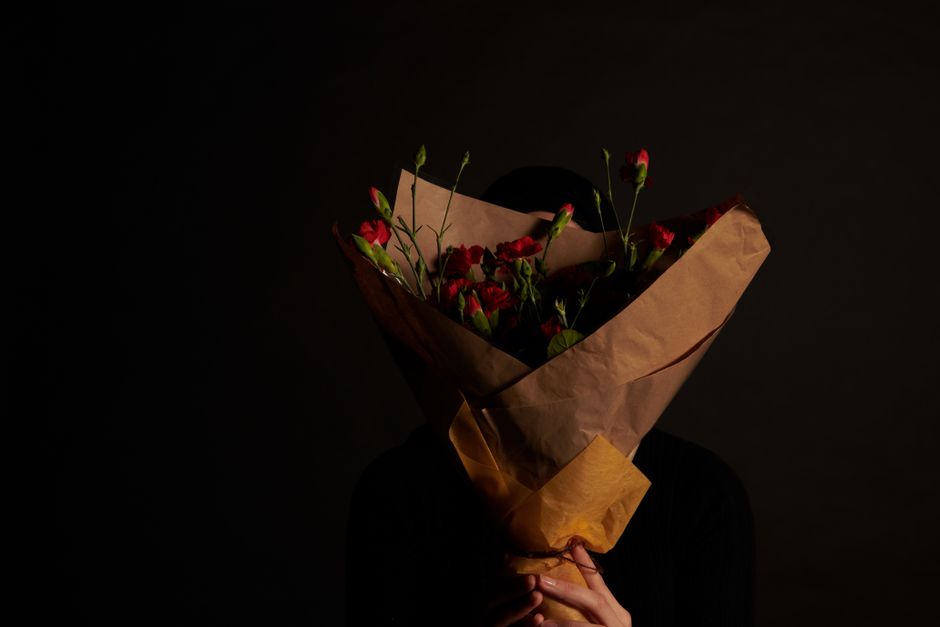 A person holding a bouquet of fresh red and pink flowers, including lisianthus and other delicate blooms, wrapped in brown kraft paper with a yellow paper band. The bouquet is positioned in front of t