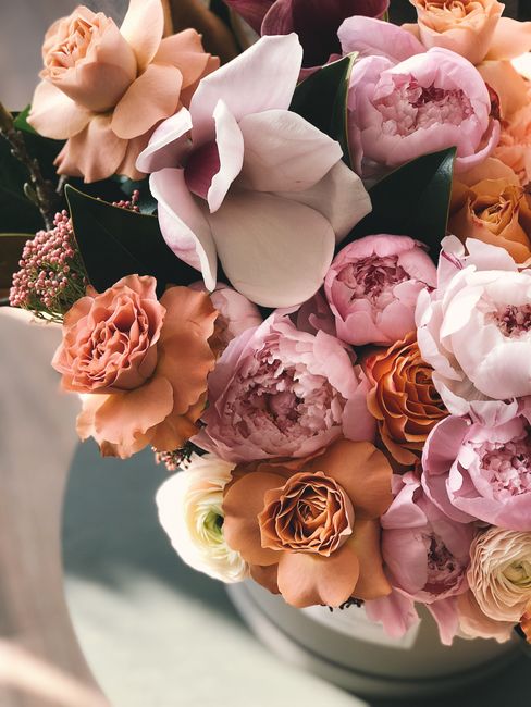 A close-up view of a floral arrangement featuring large, soft pink peonies with ruffled petals and deep pink centers, complemented by peach and apricot-colored roses and creamy white gardenias with sm