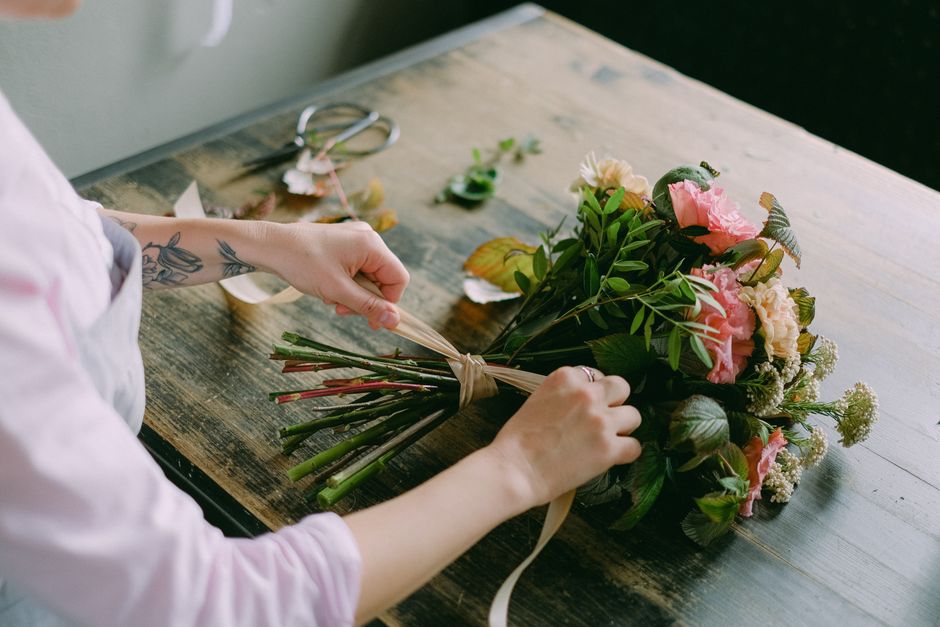 A person with tattooed arms is carefully arranging a bouquet of fresh flowers on a wooden table. The bouquet features pink peonies, creamy white roses, and green foliage tied together with a beige rib