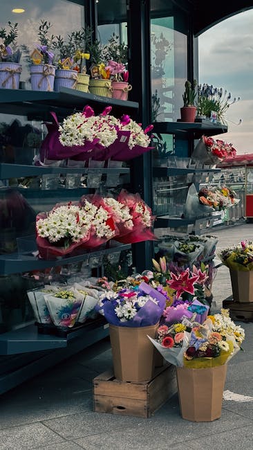 A display of various floral arrangements and bouquets inside a flower shop located at a bus stop or station. The arrangements include mixed blooms such as white chrysanthemums, pink lilies, purple ast