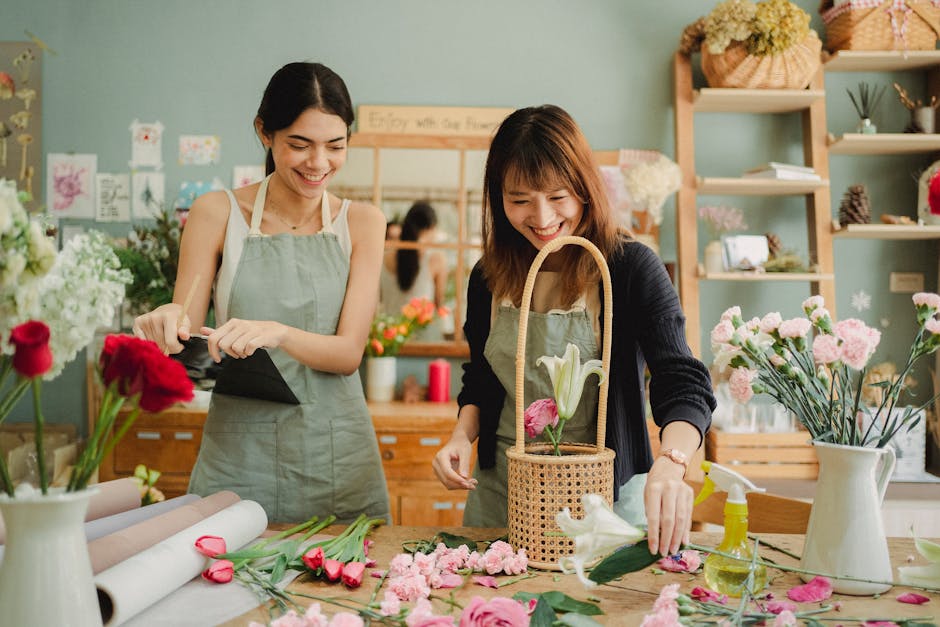 Two women working together in a bright florist shop with light green walls, arranging a bouquet of fresh flowers, including white lilies and pink roses. The woman on the left, with dark hair, is holdi