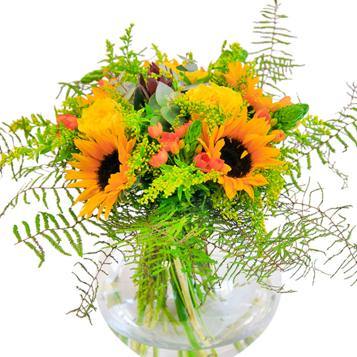 A sunlit wave of yellow and orange sweeps through this luxurious bouquet, crafted to reflect the lively yet relaxed atmosphere that Mortlake is known for. Generous sunflowers, their honeyed petals radiating in bold halos around rich, dark centers, form the visual anchor-evoking memories of summer strolls along the towpath or quiet afternoons in Barnes Common. Nestled comfortably with them, yellow garden roses reveal their full, intricate spirals, their petals lush yet refined, and their scent reminiscent of classic English gardens tucked behind the houses off Avondale Road. Intermittent dots of red Hypericum berries shimmer among the foliage, creating a contrast that sparks the entire composition to life, as if laughter and sunshine have been woven in by hand. Cascading fern fronds and pale green filler, perhaps goldenrod in its delicate bloom, lend a lively, natural movement-evoking breezy days and open fields just beyond the High Street. Threaded throughout, bluish eucalyptus leaves bring a hint of rustic wildness, balancing the structure with their soft, modern hue. All stems are gathered in a gleaming glass vase, water pure and clear, so every cut stem stands visible-showing the arrangement's freshness and allowing the natural beauty to shine. Created with skill and warmth by Flowers Mortlake, this bouquet is an inspired gift for new beginnings, cherished celebrations, or any occasion meant to brighten a home in the local SW14 neighborhood.